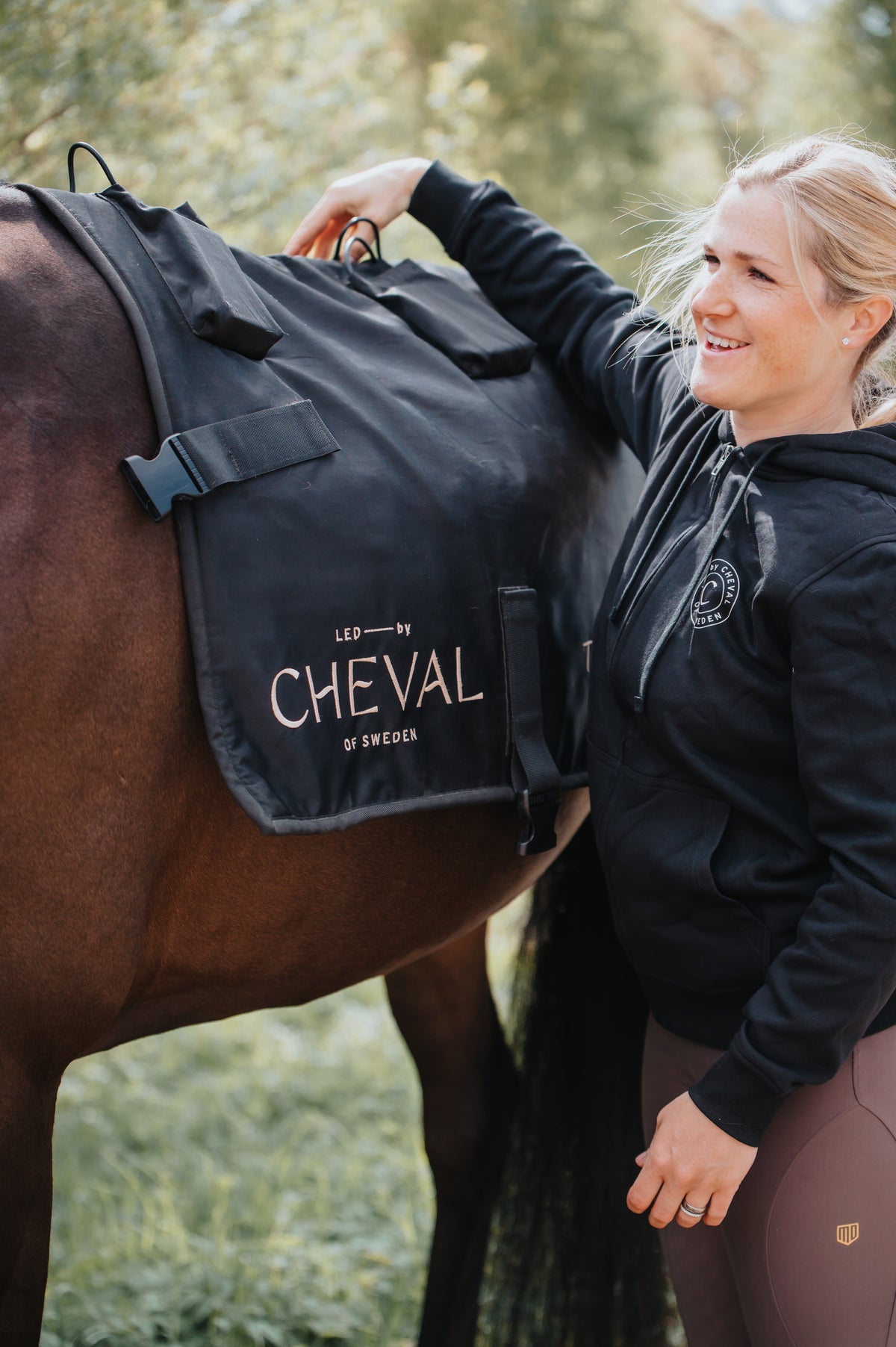 A woman settling the Sheet onto a bay horse's back outdoors, with the branded panel and buckles visible.